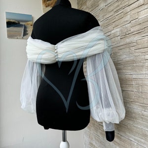 May include: White tulle bridal bolero with long, puffed sleeves and a gathered neckline. The bolero is shown on a black dress form.