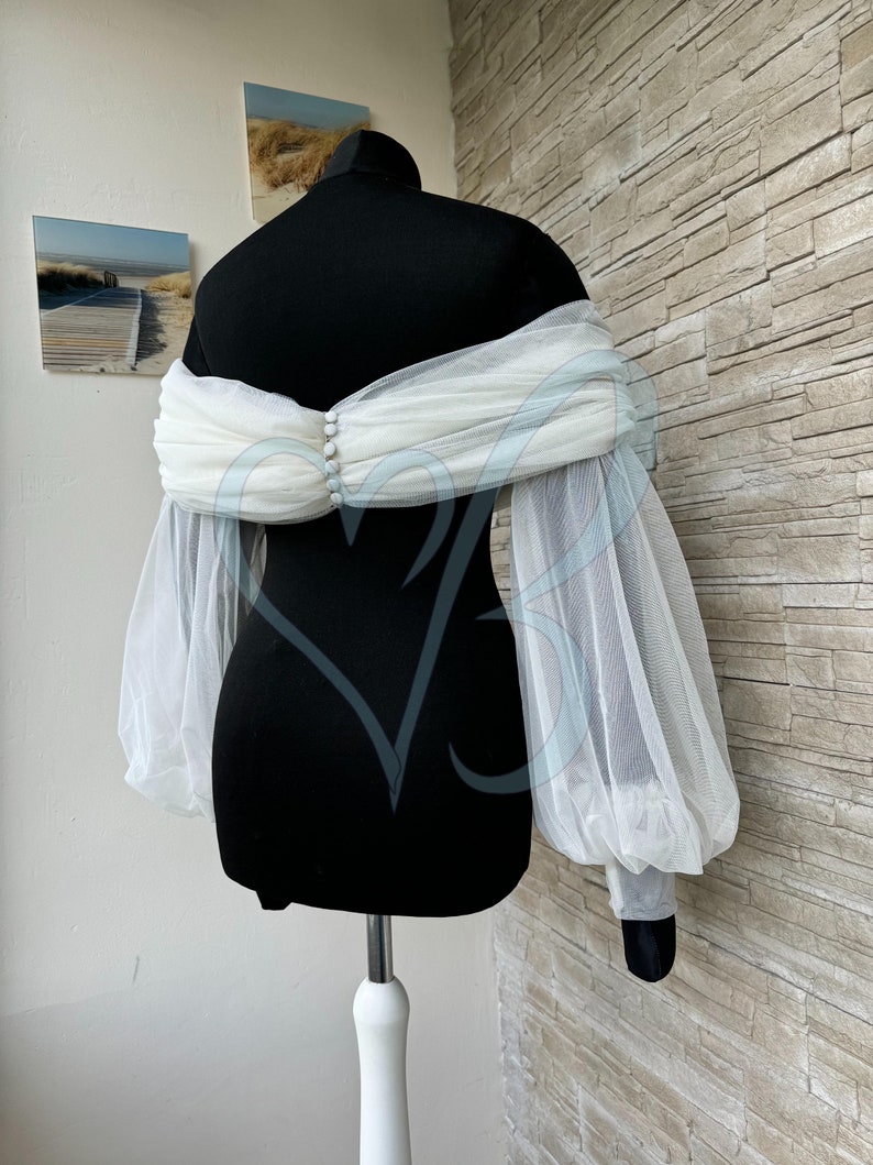 May include: White tulle bridal bolero with long, puffed sleeves and a gathered neckline. The bolero is shown on a black dress form.