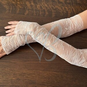 May include: A pair of sheer, white lace arm warmers. The delicate lace features a floral pattern. The arm warmers extend from the wrist to the upper arm. The background is a dark brown wooden surface.
