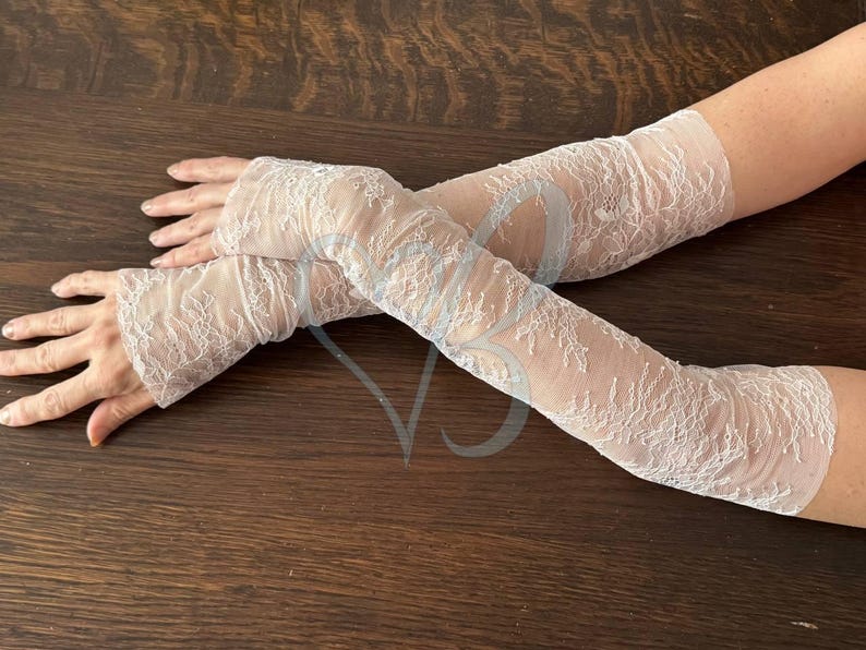 May include: A pair of sheer, white lace arm warmers. The delicate lace features a floral pattern. The arm warmers extend from the wrist to the upper arm. The background is a dark brown wooden surface.