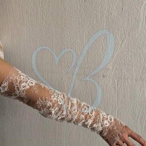 K&ouml;nnte beinhalten: Wei&szlig;e Spitzenhandschuhe mit floralen Mustern. Die Handschuhe sind lang und bedecken den Arm vom Handgelenk bis zum Oberarm. Die Spitze ist filigran und detailliert, mit einem transparenten Aussehen. Der Hintergrund ist eine neutrale, strukturierte Wand.