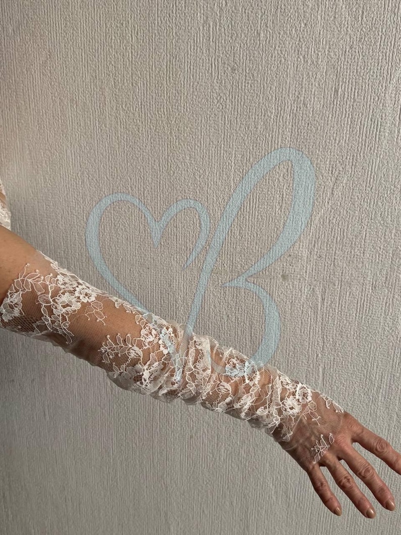 K&ouml;nnte beinhalten: Wei&szlig;e Spitzenhandschuhe mit floralen Mustern. Die Handschuhe sind lang und bedecken den Arm vom Handgelenk bis zum Oberarm. Die Spitze ist filigran und detailliert, mit einem transparenten Aussehen. Der Hintergrund ist eine neutrale, strukturierte Wand.