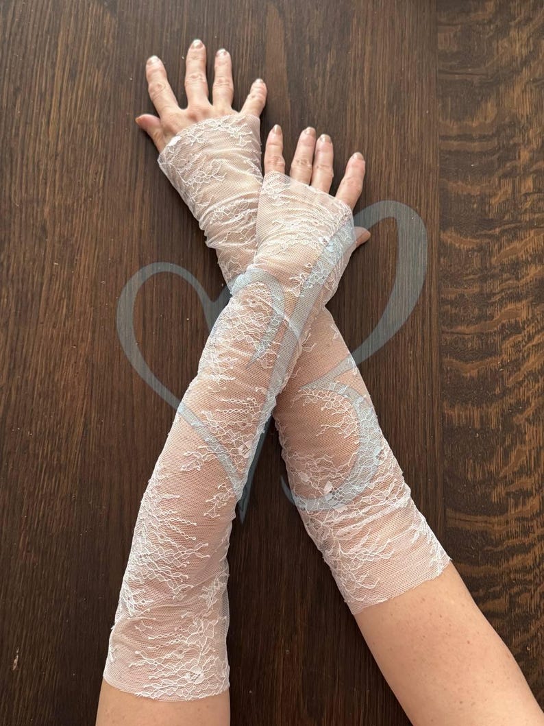 May include: A pair of sheer, off-white lace arm warmers. The delicate lace features a floral pattern and extends from the wrist to the upper arm. The arm warmers are displayed on a dark brown wooden surface.