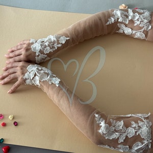 May include: A pair of white lace elbow-length gloves with a sheer mesh fabric. The gloves have a floral design and are shown on a light brown surface.