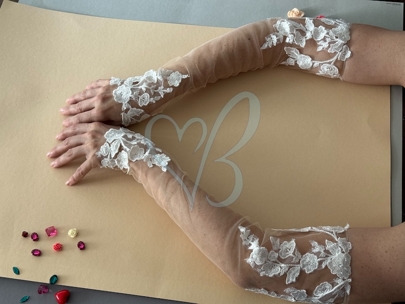 May include: A pair of white lace elbow-length gloves with a sheer mesh fabric. The gloves have a floral design and are shown on a light brown surface.