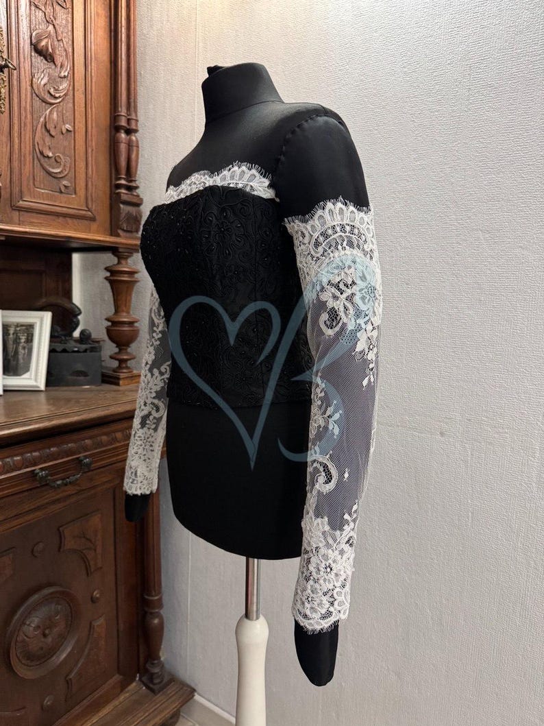 May include: A black off-the-shoulder top with long sleeves made of white lace. The top is fitted and has a delicate lace trim around the neckline.