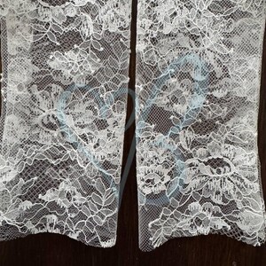 K&ouml;nnte beinhalten: Nahaufnahme von zwei Abschnitten wei&szlig;en Spitzenstoffs. Die Spitze weist ein zartes Blumenmuster mit komplizierten Details auf. Der Stoff wird wahrscheinlich f&uuml;r Brautkleider oder andere formelle Kleidungsst&uuml;cke verwendet. Der Hintergrund ist dunkel und einfarbig.