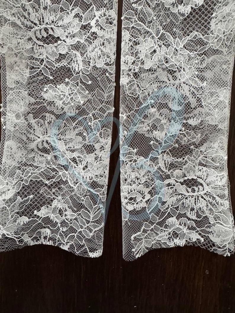 K&ouml;nnte beinhalten: Nahaufnahme von zwei Abschnitten wei&szlig;en Spitzenstoffs. Die Spitze weist ein zartes Blumenmuster mit komplizierten Details auf. Der Stoff wird wahrscheinlich f&uuml;r Brautkleider oder andere formelle Kleidungsst&uuml;cke verwendet. Der Hintergrund ist dunkel und einfarbig.