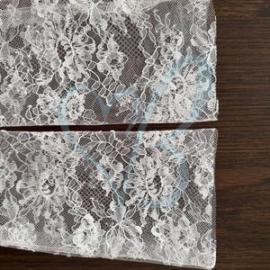 K&ouml;nnte beinhalten: Zwei St&uuml;cke wei&szlig;en floralen Spitzenstoffs. Die Spitze weist ein zartes Muster aus Blumen und Bl&auml;ttern mit dezenten blauen Akzenten auf. Der Stoff ist halbtransparent und zeigt die dunkelbraune Oberfl&auml;che darunter.