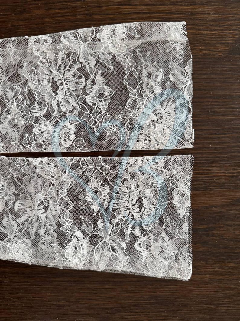 K&ouml;nnte beinhalten: Zwei St&uuml;cke wei&szlig;en floralen Spitzenstoffs. Die Spitze weist ein zartes Muster aus Blumen und Bl&auml;ttern mit dezenten blauen Akzenten auf. Der Stoff ist halbtransparent und zeigt die dunkelbraune Oberfl&auml;che darunter.