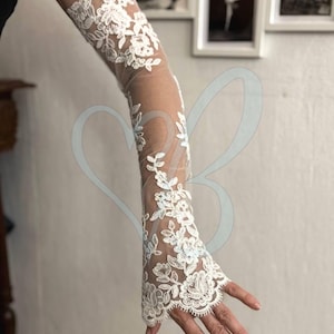 Könnte beinhalten: Ein langer, transparenter weißer Spitzenärmel, der vom Oberarm bis zur Hand reicht und mit komplizierten Blumenstickereien und gewellten Kanten versehen ist. Der zarte Netzstoff ist über die gesamte Länge mit detaillierten weißen Spitzenmustern verziert.
