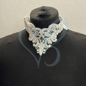 May include: A white lace collar with an intricate floral design. The collar is displayed on a black mannequin, showcasing its delicate details and elegant style. The lace is a soft white color.