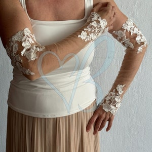 May include: A pair of white lace elbow-length gloves with a sheer mesh fabric. The gloves have a floral design and are worn on a person's arms.