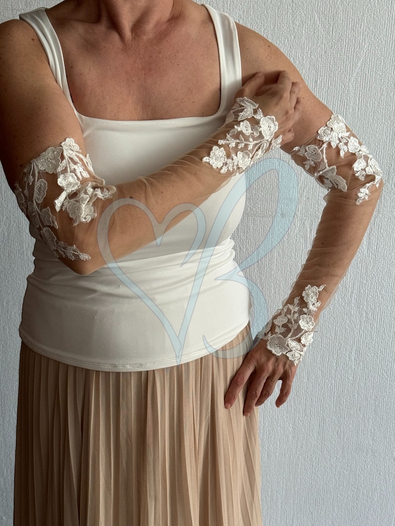 May include: A pair of white lace elbow-length gloves with a sheer mesh fabric. The gloves have a floral design and are worn on a person's arms.