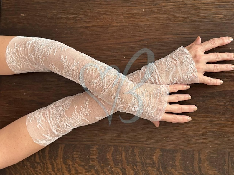 May include: A pair of white lace arm warmers with a floral design. The arm warmers are made of a sheer fabric and have a flared cuff at the wrist. The lace pattern is intricate and detailed, creating a delicate and elegant look.