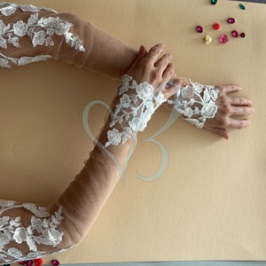 May include: A pair of white lace elbow-length gloves with a sheer mesh fabric. The gloves have a floral design and are shown on a beige background.