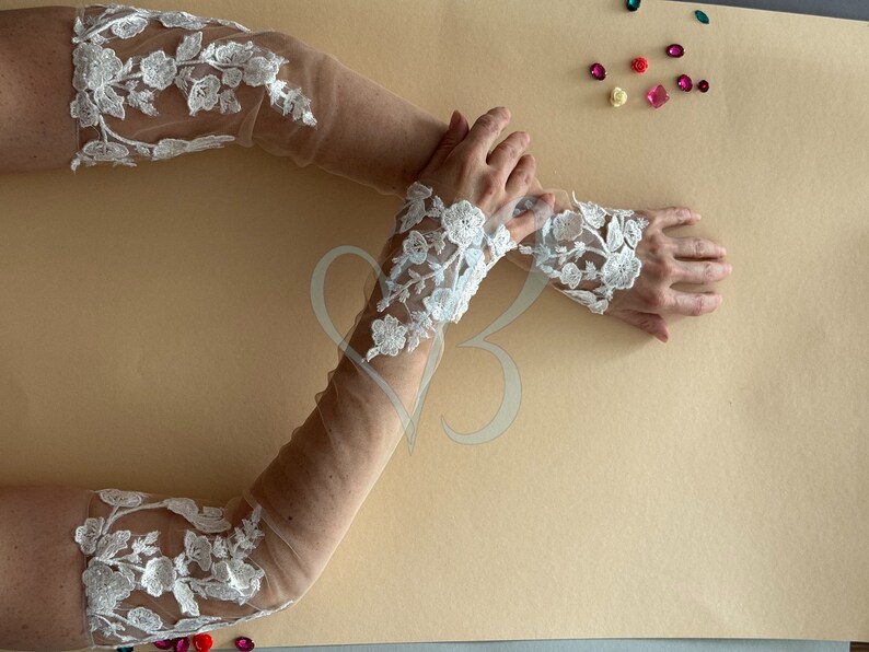 May include: A pair of white lace elbow-length gloves with a sheer mesh fabric. The gloves have a floral design and are shown on a beige background.