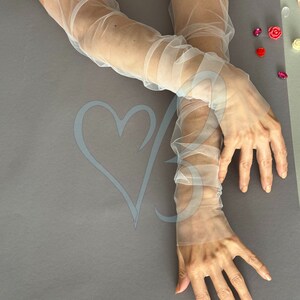 K&ouml;nnte beinhalten: Ein Paar durchsichtige wei&szlig;e T&uuml;llhandschuhe, die bis zum Ellbogen reichen. Die Handschuhe sind aus einem zarten, leichten Stoff gefertigt und haben einen leicht gerafften Look.