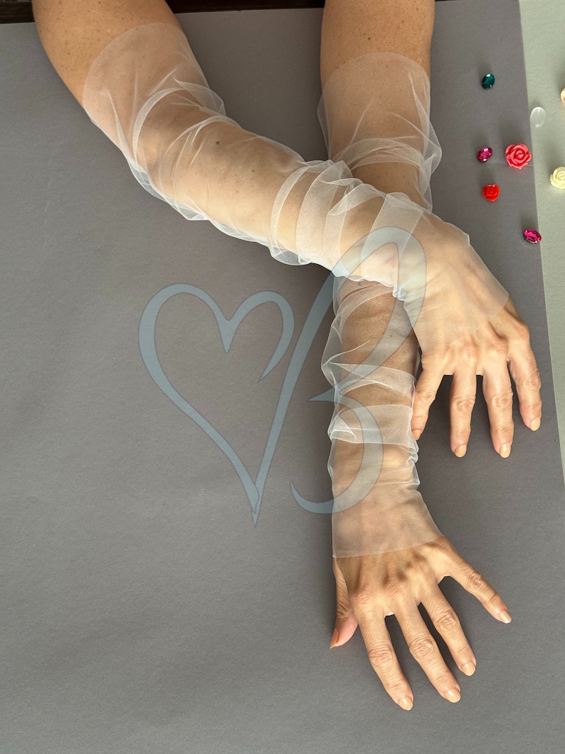K&ouml;nnte beinhalten: Ein Paar durchsichtige wei&szlig;e T&uuml;llhandschuhe, die bis zum Ellbogen reichen. Die Handschuhe sind aus einem zarten, leichten Stoff gefertigt und haben einen leicht gerafften Look.