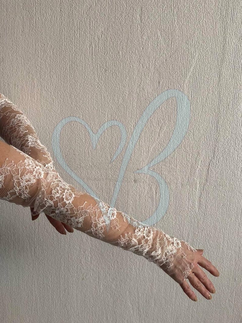 K&ouml;nnte beinhalten: Wei&szlig;e Spitzenhandschuhe mit floralem Muster. Die Handschuhe sind lang und bedecken die Arme bis zum Oberarm. Die Spitze ist detailliert und filigran, mit transparentem Hintergrund.