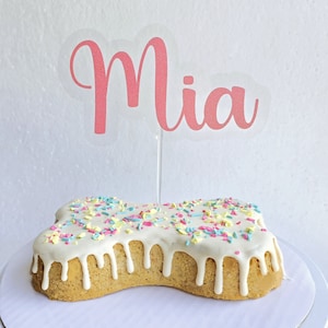 May include: A bone-shaped cake decorated with white frosting and colorful sprinkles. The cake is topped with a pink and white sign that says "Mia".