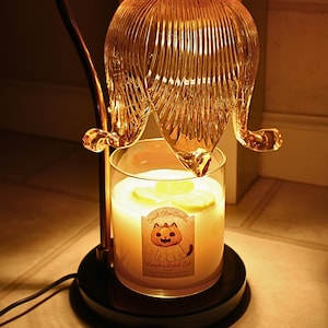 May include: A candle warmer with a glass flower-shaped shade. The candle inside has a label with a cartoon cat and the text "Pumpkin Patch Cat". The base is black and the metal arm is gold.