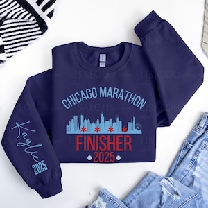 Op de afbeelding: Marineblauw sweatshirt met de tekst "Chicago Marathon Finisher 2025". Het ontwerp bevat een skyline-afbeelding en de naam "Kaylie" met het jaar 2025 op de mouw.