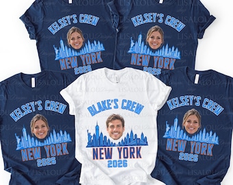 Op de afbeelding: Vier marineblauwe t-shirts en één wit t-shirt met een grafische print van de skyline van New York, een gezicht en de tekst KELSEY'S CREW of BLAKE'S CREW met NEW YORK 2026 in lichtblauwe en oranje letters op de voorzijde.