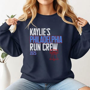 Puede incluir: Sudadera azul marino con el texto "KAYLIE'S PHILADELPHIA RUN CREW 2025" en blanco, rojo y azul. Una ilustración roja de la Campana de la Libertad está debajo. La sudadera es de estilo casual.