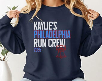 Op de afbeelding: Marineblauwe sweatshirt met de tekst "KAYLIE'S PHILADELPHIA RUN CREW 2025" in wit, rood en blauw. Een rode illustratie van de Liberty Bell staat eronder. De sweatshirt heeft een casual stijl.