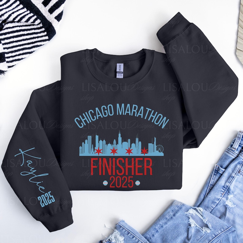 Op de afbeelding: Een marineblauw sweatshirt met de tekst "Chicago Marathon Finisher 2025". Het ontwerp bevat een skyline-afbeelding en de naam "Kaylie" op de mouw. Het sweatshirt ligt plat.