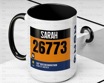 May include: White ceramic mug with a black interior and handle. The mug features a design with the name "SARAH", the number "26773", and the text "130th BOSTON MARATHON BANK OF AMERICA". The year "2026" is also printed on the mug.