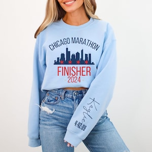 May include: A light blue crewneck sweatshirt featuring a dark blue Chicago skyline graphic with red stars, the text CHICAGO MARATHON FINISHER 2024, and a personalized name Kaylie with the time 4:42:09 printed in dark blue on the left sleeve.