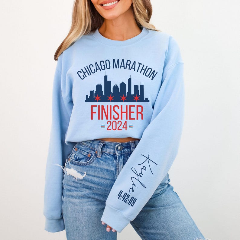 May include: A light blue crewneck sweatshirt featuring a dark blue Chicago skyline graphic with red stars, the text CHICAGO MARATHON FINISHER 2024, and a personalized name Kaylie with the time 4:42:09 printed in dark blue on the left sleeve.