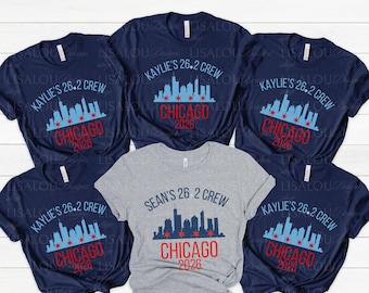 May include: Five short-sleeve t-shirts arranged in a group. Four shirts are navy blue and one is heather gray. Each shirt features a graphic of the Chicago skyline with red stars, the text "KAYLIE'S 26.2 CREW" or "SEAN'S 26.2 CREW", and "CHICAGO 2026".
