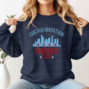 May include: A navy blue long-sleeve sweatshirt featuring a light blue Chicago skyline graphic with four red stars. The text reads CHICAGO MARATHON in light blue, FINISHER in red, and 2026 in red, flanked by two small circular icons with red and blue details.