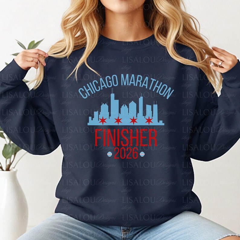 May include: A navy blue long-sleeve sweatshirt featuring a light blue Chicago skyline graphic with four red stars. The text reads CHICAGO MARATHON in light blue, FINISHER in red, and 2026 in red, flanked by two small circular icons with red and blue details.