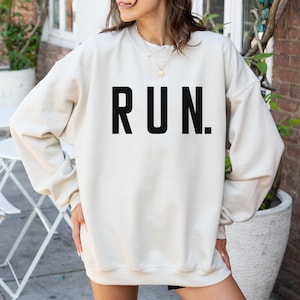 May include: A cream-colored sweatshirt with the word "RUN." printed in bold, black letters. The sweatshirt has a crew neck and long sleeves. The image shows a person wearing the sweatshirt.