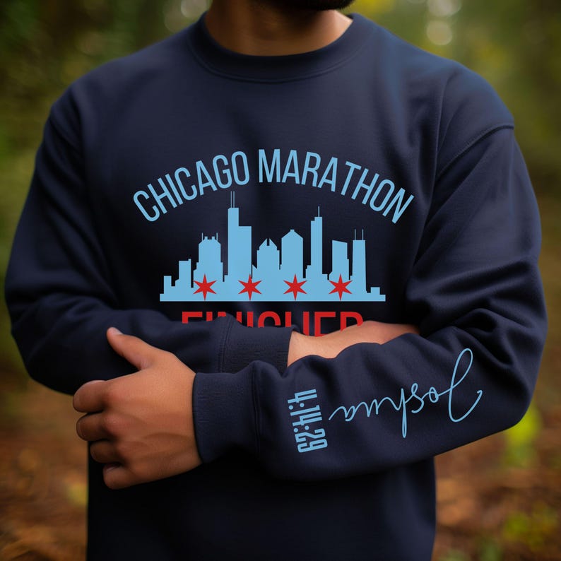 May include: A navy blue crewneck sweatshirt featuring a light blue Chicago skyline graphic with four red stars. The text reads CHICAGO MARATHON FINISHER in light blue and red. The sleeve displays the name Joshua and the time 4:14:29 in light blue.