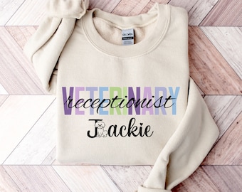 Może przedstawiać: Beżowa bluza z napisem "Veterinary receptionist Jackie" w kolorach tęczy. Obok imienia znajduje się mała ikona psa.