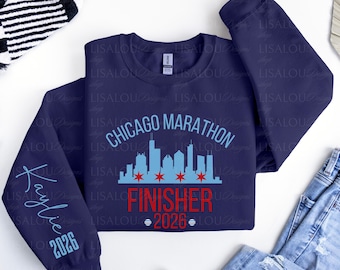 Op de afbeelding: Een marineblauw sweatshirt met ronde hals voorzien van een lichtblauwe afbeelding van de skyline van Chicago met rode sterren, de tekst "CHICAGO MARATHON FINISHER 2026" in lichtblauw en rood, en de naam "Kaylie" met "2026" op de linkermouw.