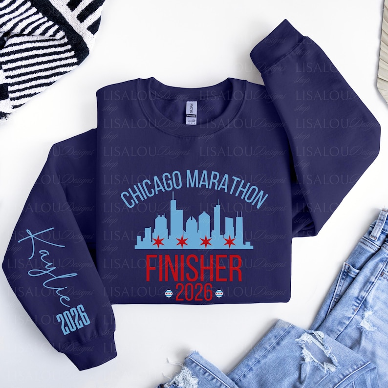 May include: A navy blue crewneck sweatshirt featuring a light blue Chicago skyline graphic with red stars, the text "CHICAGO MARATHON FINISHER 2026" in light blue and red, and the name "Kaylie" with "2026" printed in light blue on the left sleeve.