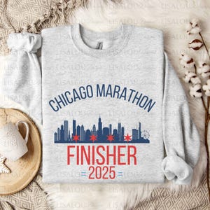 Op de afbeelding: Een lichtgrijs sweatshirt met de tekst "CHICAGO MARATHON FINISHER 2025." Het ontwerp bevat een blauwe skyline, rode sterren en de vlag van Chicago. Een comfortabele crew neck met lange mouwen.