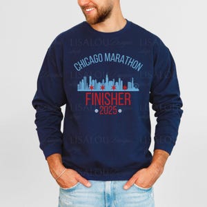 Op de afbeelding: Marineblauw sweatshirt met de tekst "Chicago Marathon Finisher 2025". Het ontwerp bevat een skyline-afbeelding met rode sterren en een blauwe rand. De sweatshirt heeft een ronde hals.