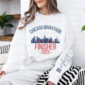 Op de afbeelding: Witte sweatshirt met de tekst "Chicago Marathon Finisher 2025" in blauw en rood. Het ontwerp bevat een skyline-afbeelding en de naam "Kaylie" op de mouw.