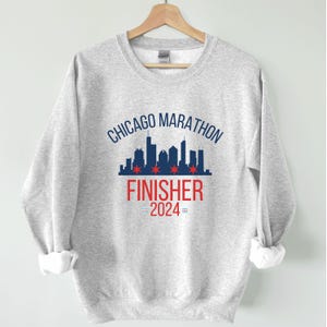 May include: A light gray crewneck sweatshirt featuring a dark blue Chicago skyline graphic with four red six-pointed stars. Below the skyline, the text reads CHICAGO MARATHON FINISHER 2024 in blue and red, with small blue and red flag icons.