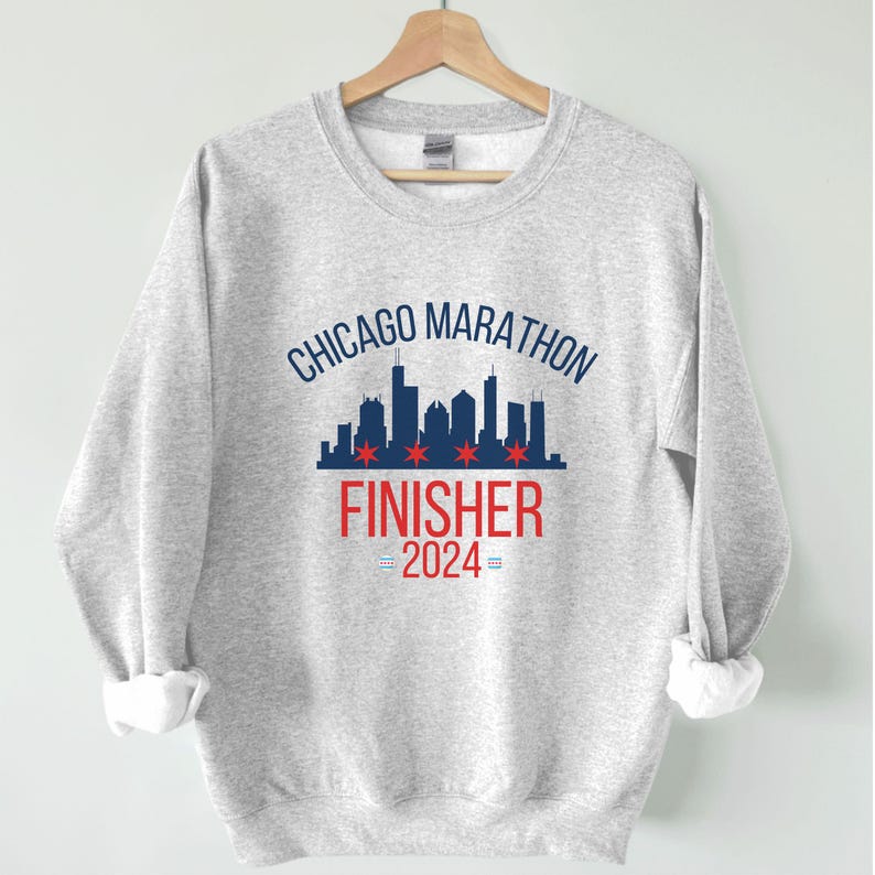 May include: A light gray crewneck sweatshirt featuring a dark blue Chicago skyline graphic with four red six-pointed stars. Below the skyline, the text reads CHICAGO MARATHON FINISHER 2024 in blue and red, with small blue and red flag icons.