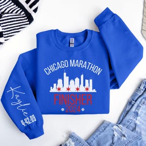 May include: A royal blue long-sleeve sweatshirt featuring a white Chicago skyline graphic with four red stars. The text reads Chicago Marathon Finisher 2024. The left sleeve is personalized with the name Kaylie and the race time 4:42:09 in white.