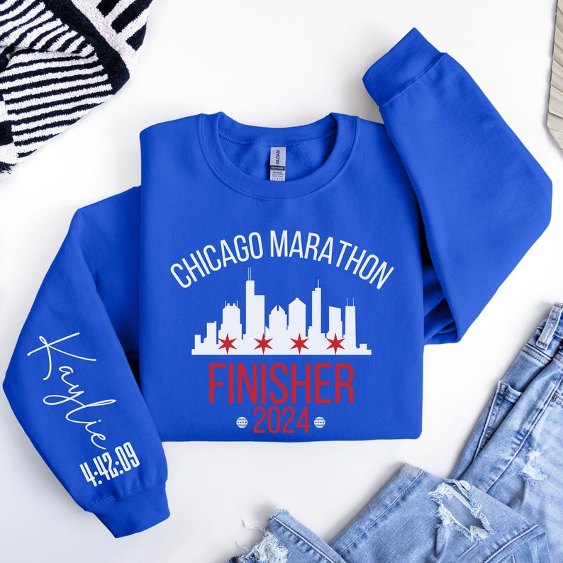 May include: A royal blue long-sleeve sweatshirt featuring a white Chicago skyline graphic with four red stars. The text reads Chicago Marathon Finisher 2024. The left sleeve is personalized with the name Kaylie and the race time 4:42:09 in white.