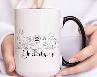 May include: White ceramic mug with a black handle. The mug has a black outline around the top rim. The mug features a black and white illustration of a stethoscope with a paw print, a dog, a cat, a rabbit, and a squirrel. The text "Dr. Johnson" is printed below the illustration.